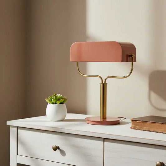 Modern table lamp with a pink shade and gold base on a white surface with a plant and book.