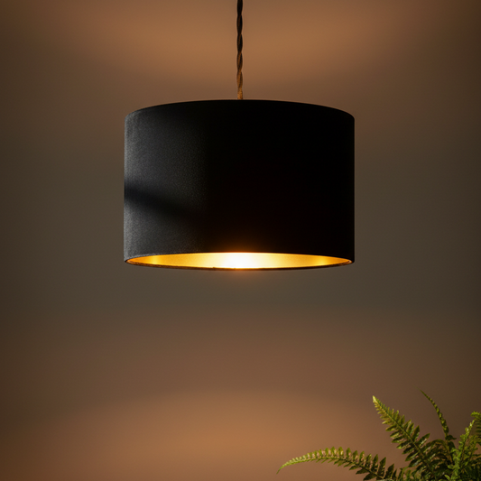 A black fabric drum pendant light with a warm gold interior, suspended by a twisted cord against a warm brown gradient background with a green fern in the lower right corner.