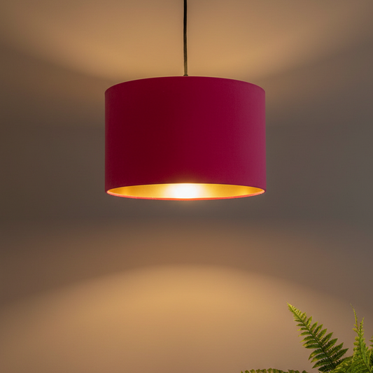A modern drum pendant light with a vibrant magenta pink fabric shade and a polished gold interior, suspended against a warm gray wall with a soft glow.