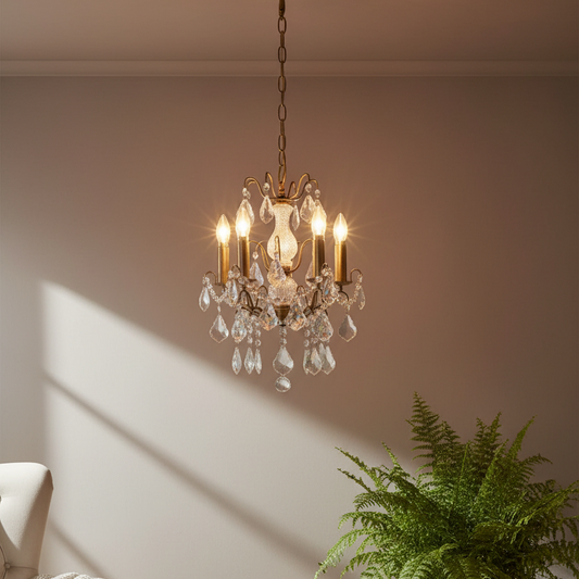A classic six-light crystal chandelier with an antique brass finish hangs from a chain in a living room setting, casting warm light on a beige wall next to a green fern plant.