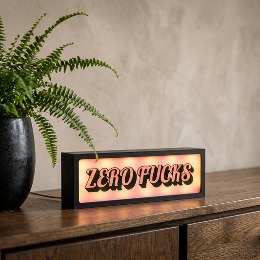 A rectangular black LED sign with the words 'ZERO FUCKS' in raised red letters, glowing with a warm light, placed on a wooden cabinet next to a potted fern against a textured beige wall.