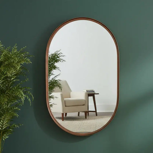 Oval mirror with wooden frame on a green wall reflecting a room with a chair and table.