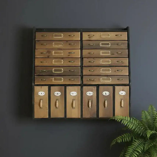Wooden drawer unit with brass handles against a dark gray wall, accompanied by a potted fern.