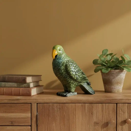 Green parrot figurine on a wooden surface with books and a plant against a beige wall.