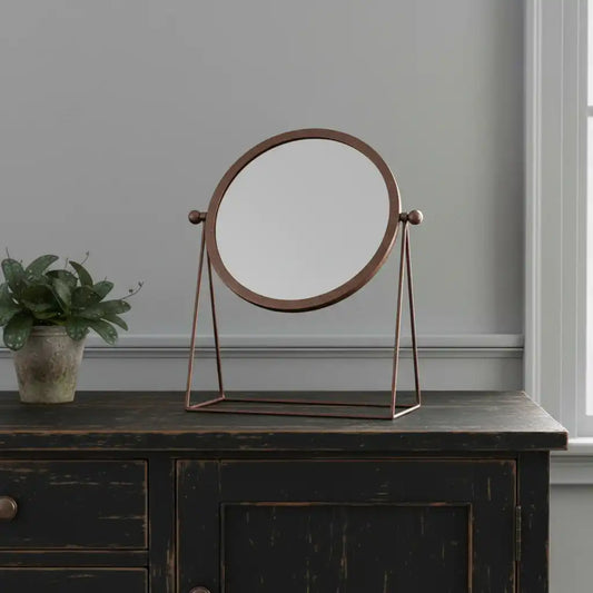 Round stand-up mirror with bronze frame on a dark wooden surface, with a plant beside it.