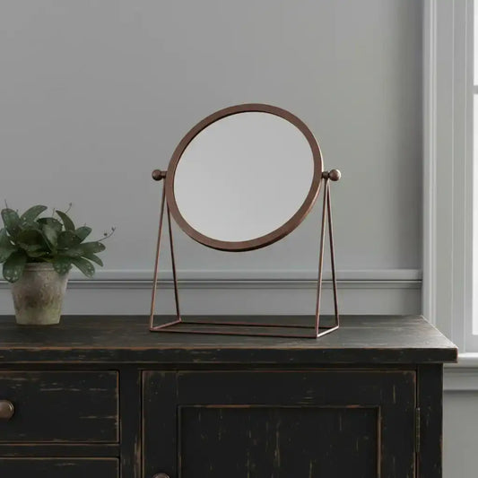 Round stand-up mirror with bronze frame on a dark wooden surface, with a plant beside it.