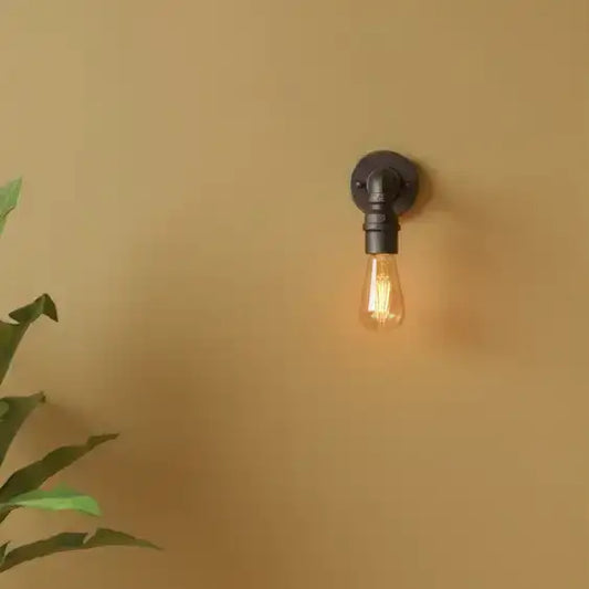 Wall-mounted light fixture with a lit bulb on a beige wall, featuring a plant on the left.
