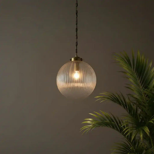 Glass pendant light with gold fixture hanging against a dark background with a plant to the right.