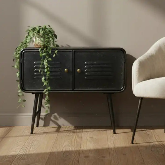 Black side table with gold handles next to a beige armchair and plant on a wooden floor.