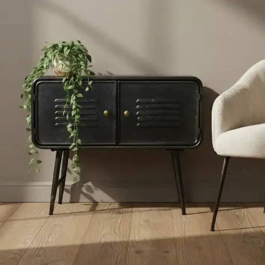 Black side table with gold handles next to a beige armchair and plant on a wooden floor.