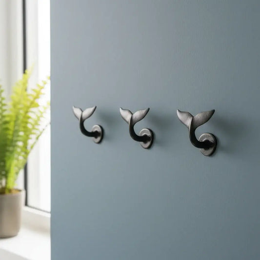Three whale tail-shaped wall hooks on a gray wall with a plant in the corner.