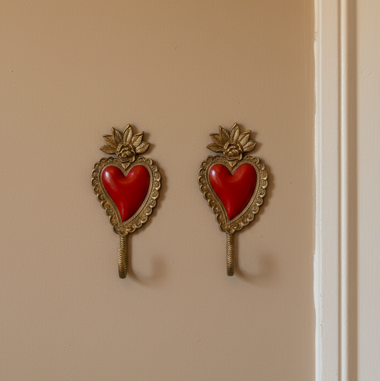 A pair of vintage-style wall hooks shaped like sacred hearts, featuring red enamel hearts set in ornate gold-tone metal frames, mounted on a beige wall.