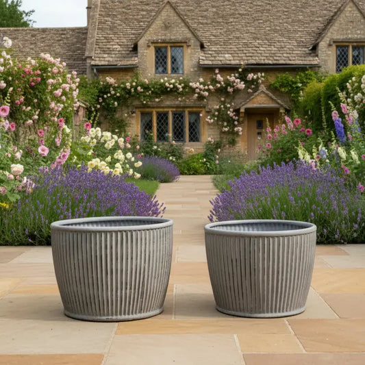 Two gray planters in front of a charming house with blooming flowers.