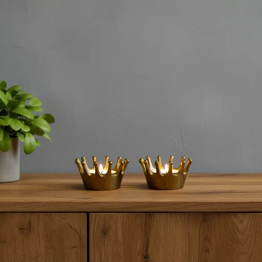Two gold crown-shaped candle holders on a wooden surface with a plant in the background.