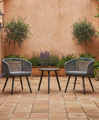 Two outdoor chairs with a small table on a tiled patio with plants and a wall in the background.