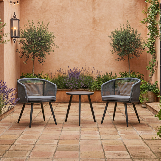 Two outdoor chairs with a small table on a tiled patio with plants and a wall in the background.