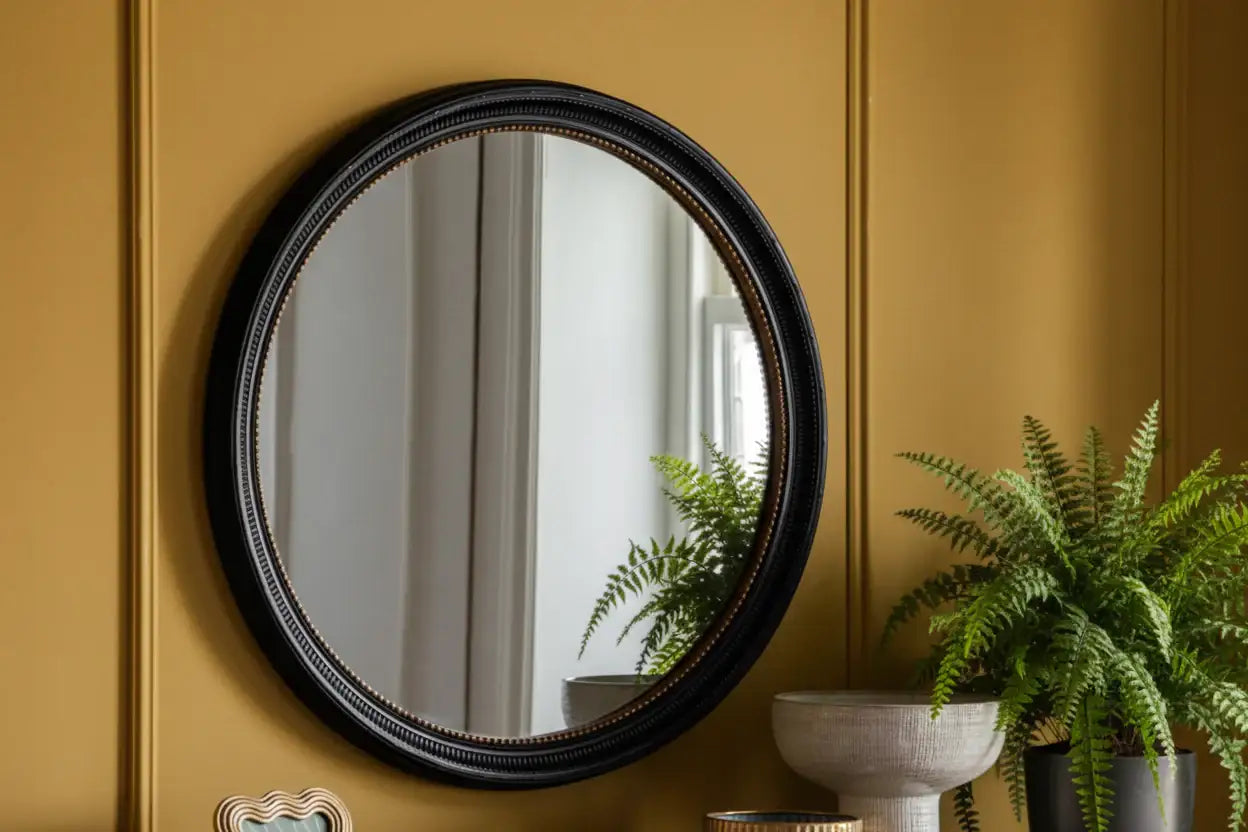 Round mirror with black frame on a yellow wall with a plant in the background