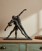 Statue of two dancers on a wooden surface with a plant and book in the background