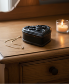 A black heart-shaped ornate jewelry box with a gold necklace resting beside it on a wooden nightstand, with a lit candle in the background.
