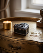 A small black ornate jewelry box with a heart-shaped lid sits on a distressed bronze-colored wooden table next to a lit gold candle and a gold chain necklace.