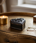 A small black ornate jewelry box with a carved lid sits on a distressed gold-toned wooden table next to a lit gold candle and a gold chain bracelet.