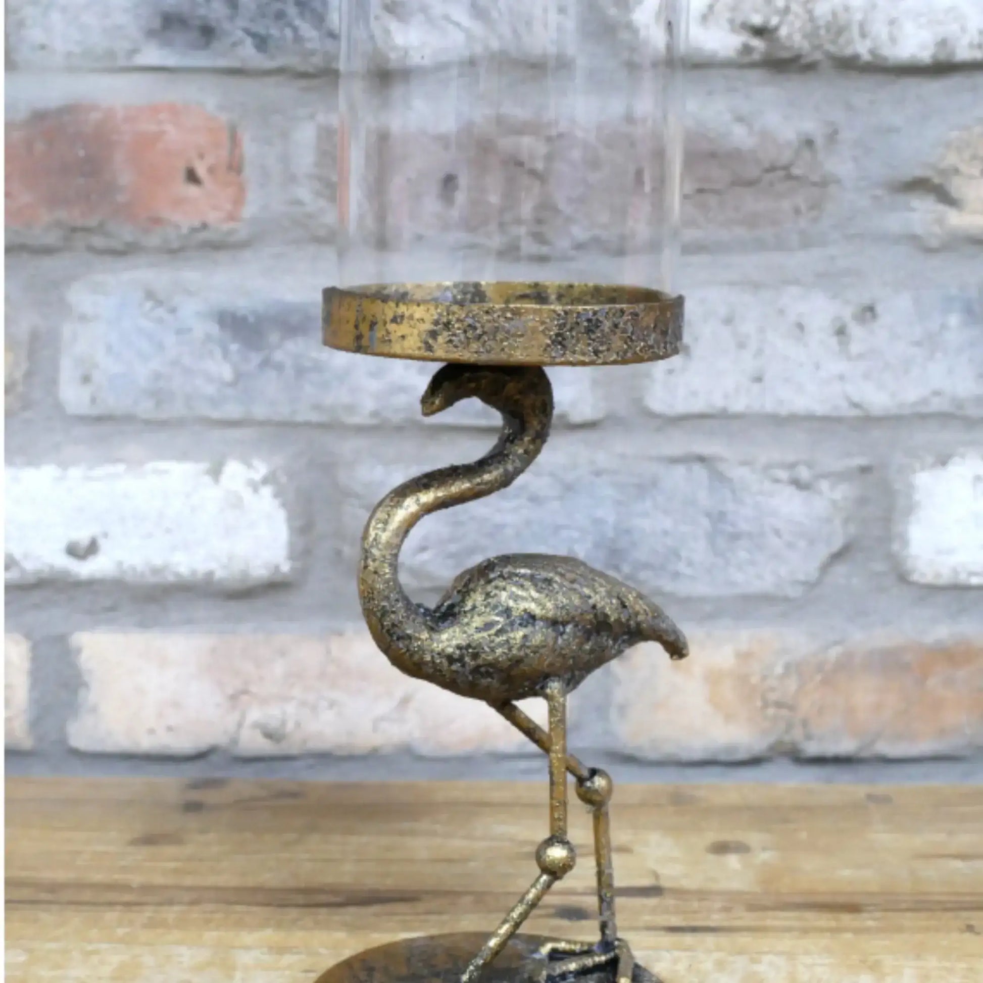 Decorative metal flamingo candle holder with a glass cover against a stone wall background.
