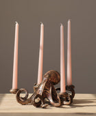 An octopus-shaped metal candle holder with four slim pink taper candles, displayed on a light wood surface against a dark gray wall with a green plant leaf in the background.