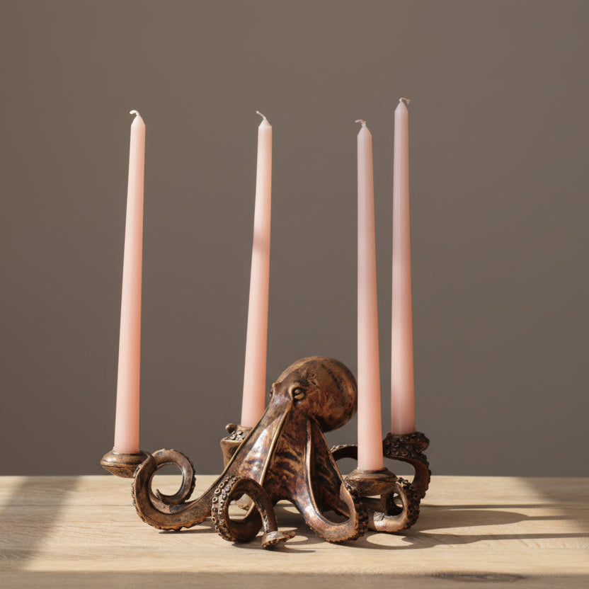An octopus-shaped metal candle holder with four slim pink taper candles, displayed on a light wood surface against a dark gray wall with a green plant leaf in the background.