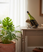 A green wine bottle with a black label and gold script stands on a decorative metallic octopus sculpture on a rustic wooden table. The scene is styled in a warm, sunlit room with a potted monstera plant in the foreground and a beige wall in the background.