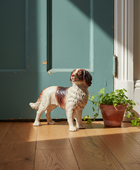 A ceramic dog figurine standing on a wooden floor next to a potted plant, positioned in front of a teal door with warm sunlight casting dramatic shadows.
