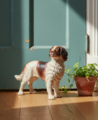 A ceramic dog figurine in a white and brown spaniel style stands on a wooden floor beside a potted green plant in front of a teal door.