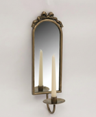 An antique-style bronze wall sconce with an arched mirror and candle-shaped lights reflected in the glass, set against a plain light gray wall.