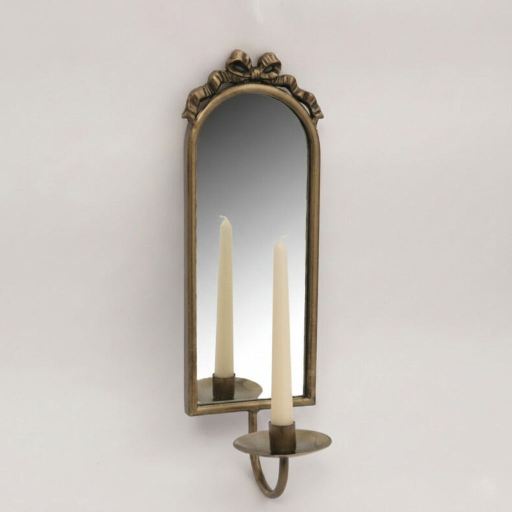 An antique-style bronze wall sconce with an arched mirror and candle-shaped lights reflected in the glass, set against a plain light gray wall.