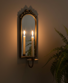 An ornate arched wall mirror with a gold frame and candle-style sconces holding lit white candles, mounted on a warm beige wall with a green fern plant visible on the right.