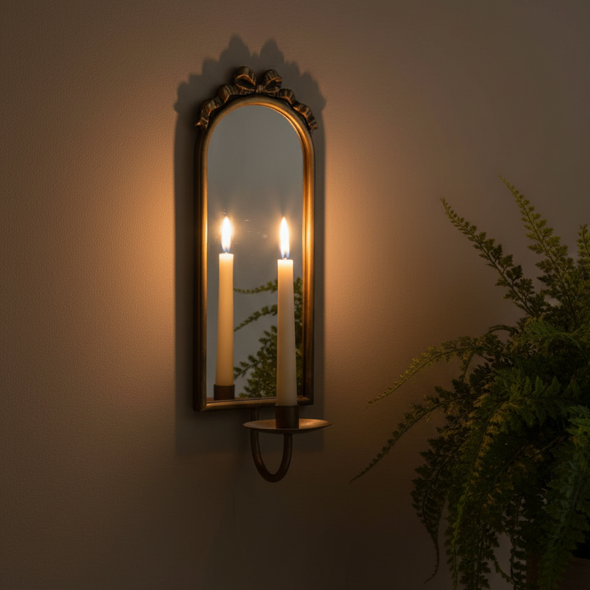 An ornate arched wall mirror with a gold frame and candle-style sconces holding lit white candles, mounted on a warm beige wall with a green fern plant visible on the right.