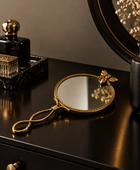 A decorative handheld mirror with an ornate gold frame, a bee accent at the top, and a loop handle, styled on a dark wooden dresser next to perfume bottles.