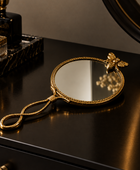An ornate gold handheld mirror with a decorative handle and a small bee ornament at the top, displayed on a dark wooden vanity table.