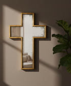 Decorative cross-shaped mirror with gold frame on a wall with a plant to the right.
