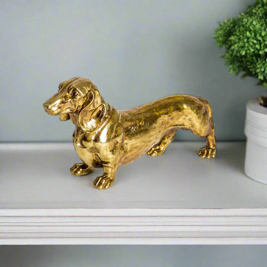Gold dachshund figurine on a white surface with a light gray wall and green plant in the background