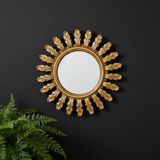 Decorative gold sunburst mirror on a black wall with a plant in the foreground.