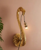 Gold flamingo-shaped wall lamp on a pink wall with a plant to the left.