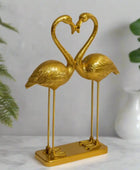 Gold flamingo sculpture forming a heart shape on a white surface with green plants in the background