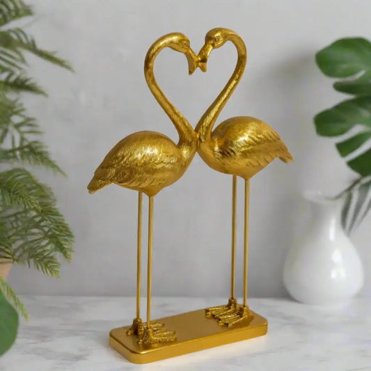 Gold flamingo sculpture forming a heart shape on a white surface with green plants in the background