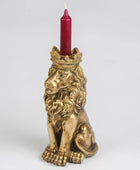 A gold-toned lion figurine candle holder with a crown on its head, holding a red taper candle, photographed against a light gray studio background.