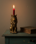 A red candle in a gold lion-shaped holder with a crown sits on a distressed teal table next to an old book against a textured beige wall.