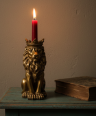 A brass lion candle holder with a crown, holding a lit red taper candle, displayed on a distressed teal wooden table next to an old book against a textured beige wall.