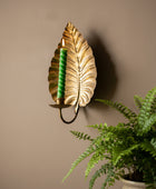 A decorative wall-mounted leaf-shaped candle holder in a brushed gold finish, holding a black braided candle, set against a dark gray background with a soft shadow and blurred green leaves at the bottom right.