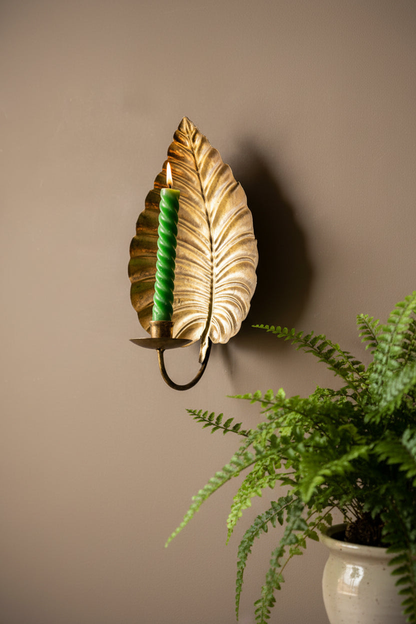 A decorative wall-mounted leaf-shaped candle holder in a brushed gold finish, holding a black braided candle, set against a dark gray background with a soft shadow and blurred green leaves at the bottom right.