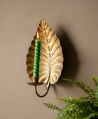 A decorative wall sconce shaped like a metallic gold leaf, holding a lit green twisted candle, mounted on a warm beige wall with a green fern in the foreground.