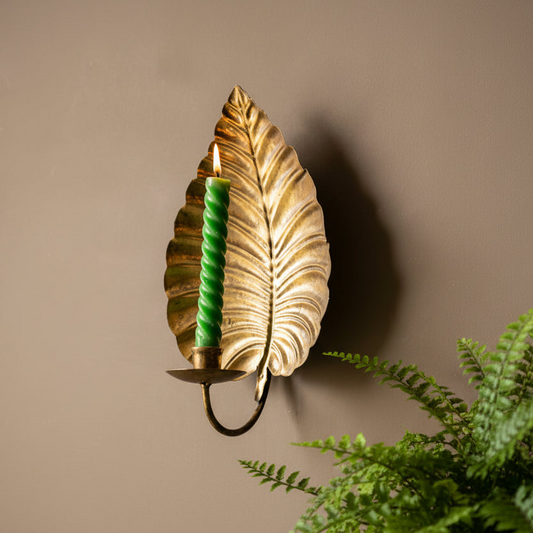 A decorative wall sconce shaped like a metallic gold leaf, holding a lit green twisted candle, mounted on a warm beige wall with a green fern in the foreground.